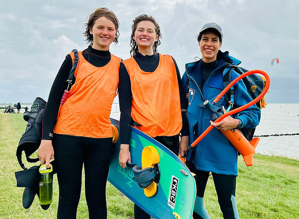 Drei lächelnde Kitesurferinnen mit Kitesurf- und Holland007-Ausrüstung am Strand, Kiteboarding Wassersport Reiseziel