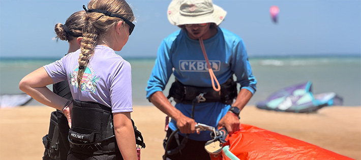 Kitekurs Kids: Kitesurfing instructeur leert kind kiteboarding uitrusting op strand tijdens watersport les