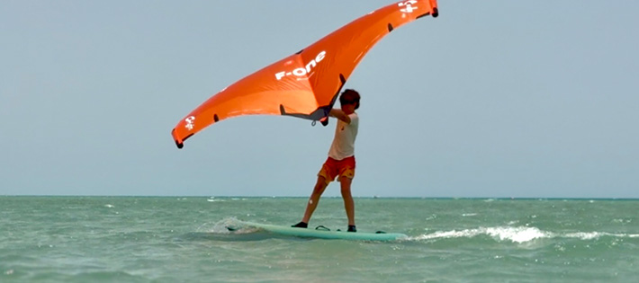 Wingfoilkurs Kids - Jovem praticando wingfoil no mar turquesa com asa laranja, kitesurf esportes aquáticos praia