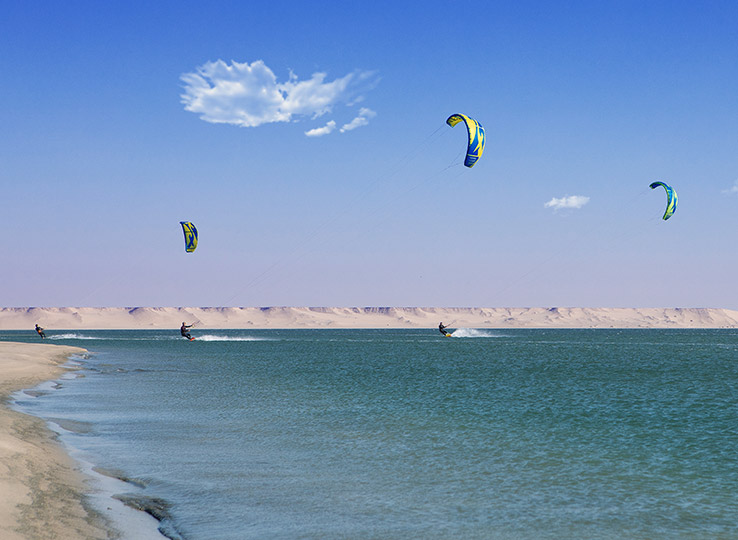 Speedspot kitesurfen op turquoise strand met drie kitesurfers en kleurrijke kites tegen woestijnachtergrond
