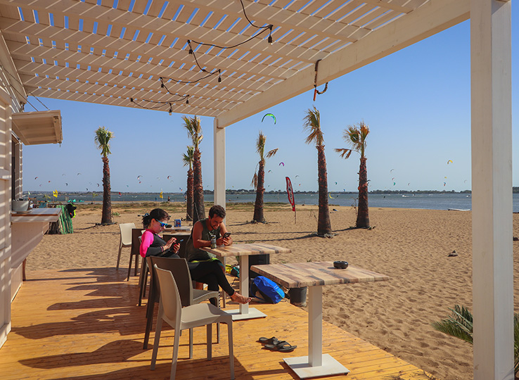 Sizilien Kitecenter beachfront café with guests relaxing under pergola, palm trees and kitesurfing beach in background