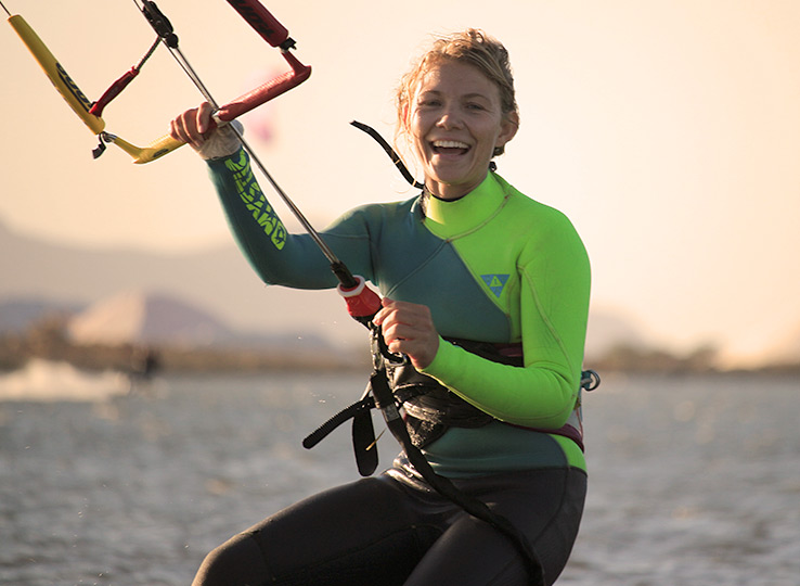 Smiling female kitesurfer in wetsuit holding kite control bar at beach - kiteboarding water sports destination