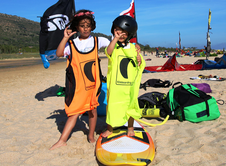 Twee kinderen in kitesurfing vesten op strand met kite uitrusting en vlaggen - kiteles voor kinderen op strand